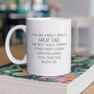 May include: White ceramic mug with a black printed message: "YOU ARE A REALLY. REALLY GREAT DAD. THE BEST. REALLY TERRIFIC. OTHER DADS? LOSERS! EVERYONE AGREES. TOTAL DISASTERS. BELIEVE ME." The mug has a curved handle.