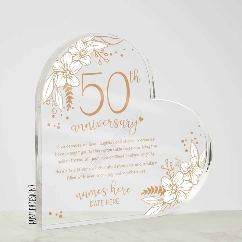Puede incluir: Una placa acr&iacute;lica transparente en forma de coraz&oacute;n con dise&ntilde;os florales dorados y el texto "50 aniversario". La placa tambi&eacute;n incluye el texto "Cinco d&eacute;cadas de amor, risas y recuerdos compartidos te han llevado a este hito extraordinario. Que el hilo dorado de tu amor siga brillando. Brindo por una vida de momentos preciados y un futuro lleno de a&uacute;n m&aacute;s alegr&iacute;a y uni&oacute;n. nombres aqu&iacute; FECHA AQU&Iacute;".