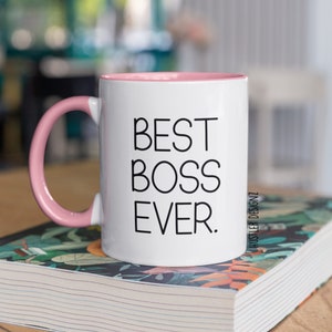 May include: White ceramic mug with a pink rim and handle. The mug has black text that reads "BEST BOSS EVER." The text "HUSTLER DESIGNZ" is printed in smaller black text on the right side of the mug.