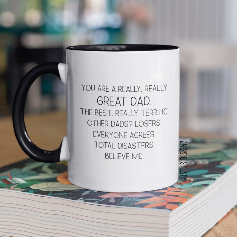 May include: White ceramic mug with a black handle and interior rim. The mug features the text "YOU ARE A REALLY. REALLY GREAT DAD." and other humorous phrases. The mug is sitting on a book.