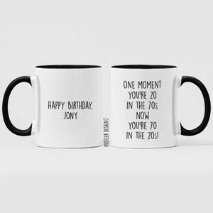 Może przedstawiać: Dwa białe kubki do kawy z czarnymi uchwytami i czarnymi obw&oacute;dkami. Kubek po lewej stronie ma napis "Happy Birthday, Jony". Kubek po prawej stronie ma napis "One moment you're 20 in the 70s. Now you're 70 in the 20s!"