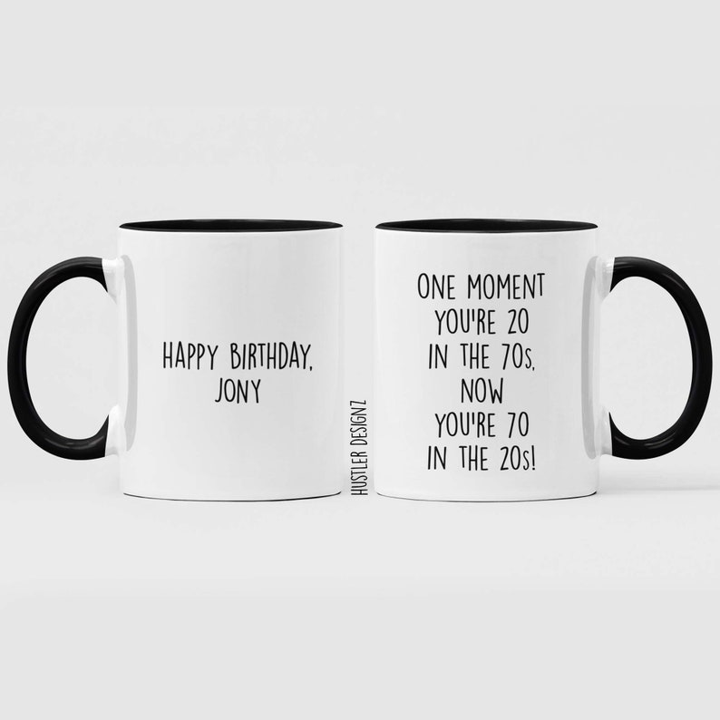 Może przedstawiać: Dwa białe kubki do kawy z czarnymi uchwytami i czarnymi obw&oacute;dkami. Kubek po lewej stronie ma napis "Happy Birthday, Jony". Kubek po prawej stronie ma napis "One moment you're 20 in the 70s. Now you're 70 in the 20s!"