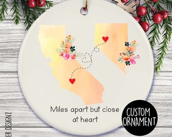 Puede incluir: Adorno de cerámica blanca con ilustraciones de acuarela de California y Nevada, conectadas por una línea punteada con corazones. Arreglos florales adornan cada estado. El texto dice "Miles apart but close at heart."