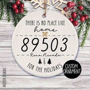 May include: A round white ceramic Christmas ornament featuring the black text There is no place like home, the zip code 89503, Reno, Nevada, and For the holidays. The design includes gold bell icons, three small black pine tree silhouettes, and a gold string.