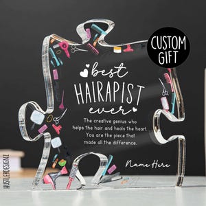 Puede incluir: Una pieza de rompecabezas de acrílico transparente con forma de corazón con un tema de salón de belleza. La pieza del rompecabezas tiene un fondo blanco con el texto "best HAIRAPIST ever" y un pequeño corazón a cada lado del texto. La pieza del rompecabezas también tiene un pequeño corazón en la esquina inferior derecha con el texto "The creative genius who helps the hair and heals the heart. You are the piece that made all the difference." La pieza del rompecabezas tiene un espacio en blanco en la esquina inferior derecha para un nombre. La pieza del rompecabezas está sobre un fondo negro. El texto "CUSTOM GIFT" está en la esquina superior derecha de la imagen.