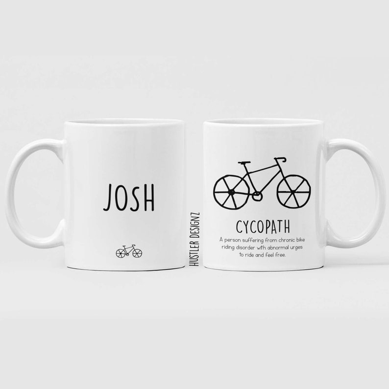 May include: Two white ceramic mugs with black text and a bicycle illustration. The mug on the left says "JOSH" and the mug on the right says "CYCOPATH" and defines it as "A person suffering from chronic bike riding disorder with abnormal urges to ride and feel free."