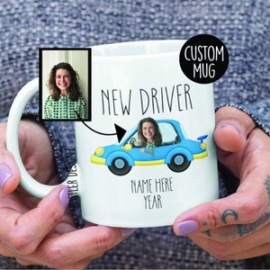 May include: White ceramic mug with a blue car illustration and the text "NEW DRIVER". A person's face is in the driver's seat of the car. The text "NAME HERE YEAR" is below the car. The mug has a custom mug label.