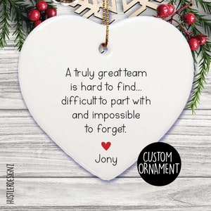 Puede incluir: Un adorno de cerámica blanco en forma de corazón con el texto impreso en negro: A truly great team is hard to find... difficult to part with and impossible to forget. Debajo del texto hay un pequeño corazón rojo y el nombre Jony, con un cordón dorado.