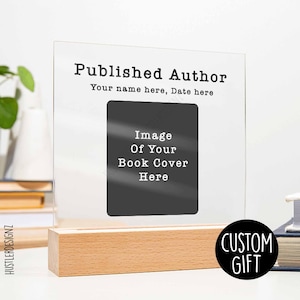 Op de afbeelding: Een helder acryl frame met een houten basis, waarop een zwart-witte afbeelding van een boekomslag te zien is. Het frame heeft de tekst "Published Author" en "Your name here, Date here". De tekst "CUSTOM GIFT" is zichtbaar in een zwarte cirkel.