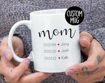 Könnte beinhalten: Weiße Keramiktasse mit dem Wort "mom" in schwarzer Kursivschrift. Unter dem Wort "mom" stehen drei Zeilen Text: "01.01.99 ♥ Jony", "01.01.20 ♥ Josh" und "01.01.22 ♥ Kate".