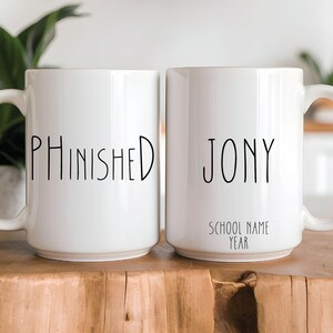May include: Two white ceramic mugs with black text. One mug reads "PHINISHED", the other reads "JONY" with "SCHOOL NAME YEAR" below. Both mugs have a handle and are on a wooden surface.
