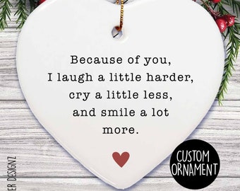 May include: A white heart-shaped ornament with the text "Because of you, I laugh a little harder, cry a little less, and smile a lot more." A small red heart is at the bottom. The ornament has a gold string for hanging and the words "CUSTOM ORNAMENT" in a black circle.