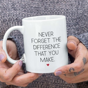 May include: White ceramic mug with the text "NEVER FORGET THE DIFFERENCE THAT YOU MAKE" in black, with a small red heart. The mug has a curved handle and is held by a person.
