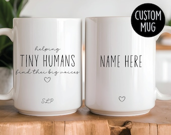 Op de afbeelding: Twee witte keramische mokken met zwarte tekst. Eén mok zegt "helping tiny humans find their big voices" met een hartje en "SLP". De andere mok zegt "NAME HERE" met een hartje. De mokken staan op een houten oppervlak.