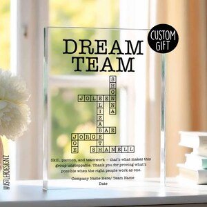 以下が含まれることがあります： 太字の黒い文字で「DREAM TEAM」と書かれた透明なアクリル板。チームメンバーの名前はクロスワードパズルのように配置されています。「CUSTOM GIFT」というテキストと、チームワークに関するモチベーションを上げる引用句が含まれています。