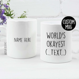 Op de afbeelding: Twee witte keramische mokken. Eén mok heeft de tekst "NAME HERE" in het zwart. De andere mok heeft de tekst "WORLD'S OKAYEST (.TEXT..)" in het zwart. Een zwarte cirkel met de woorden "CUSTOM MUG" staat op de rechter mok. De mokken staan op een wit oppervlak.
