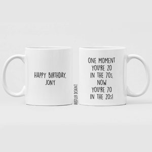 Może przedstawiać: Dwa białe ceramiczne kubki z czarnym tekstem. Kubek po lewej stronie ma napis "Happy Birthday, Jony". Kubek po prawej stronie ma napis "One moment you're 20 in the 70s. Now you're 70 in the 20s!"