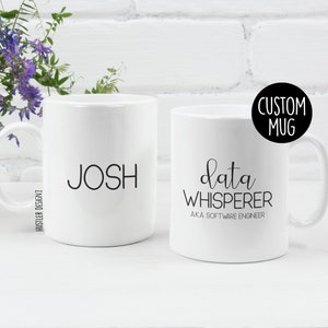 Puede incluir: Dos tazas de cerámica blanca. Una taza tiene el nombre "JOSH" impreso en ella. La otra taza tiene el texto "data WHISPERER AKA SOFTWARE ENGINEER" impreso en ella. Las tazas están sobre una superficie blanca.