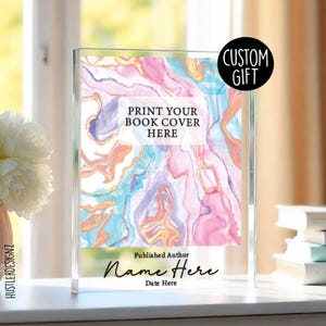 May include: A clear acrylic book cover display featuring a vibrant abstract design in pink, blue, and gold. The text "PRINT YOUR BOOK COVER HERE" is displayed, with space for a name and date. A black circle with "CUSTOM GIFT" is in the upper right corner.