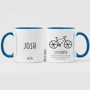 May include: White ceramic mug with a blue rim and handle. The mug has a black bicycle illustration and the text "CYCOPATH" with a definition below. The other side of the mug has the name "JOSH" and a small bicycle illustration.