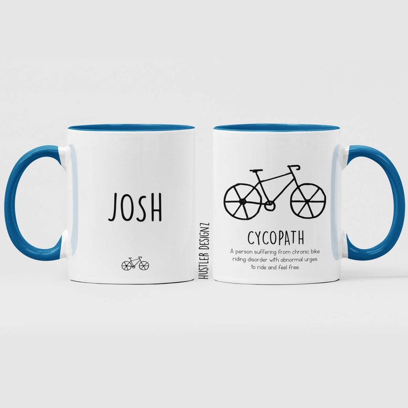 May include: White ceramic mug with a blue rim and handle. The mug has a black bicycle illustration and the text "CYCOPATH" with a definition below. The other side of the mug has the name "JOSH" and a small bicycle illustration.
