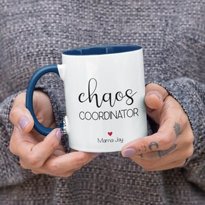 May include: White ceramic coffee mug with a blue rim and handle. The mug has black text that reads "chaos coordinator" and "Mama Jay" with a small red heart.