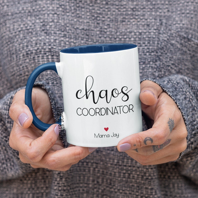 May include: White ceramic coffee mug with a blue rim and handle. The mug has black text that reads "chaos coordinator" and "Mama Jay" with a small red heart.