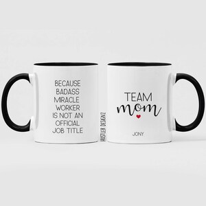 May include: Two white coffee mugs with black handles and black rims. The mug on the left has the text "Because badass miracle worker is not an official job title". The mug on the right has the text "Team mom" with a small red heart and the name "Jony" underneath.