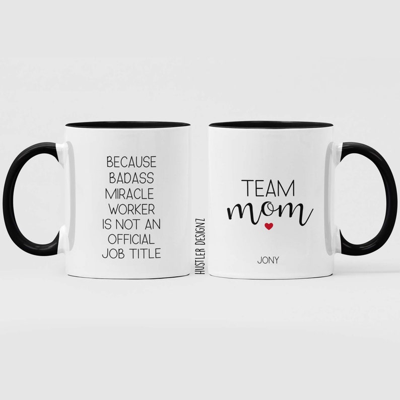 May include: Two white coffee mugs with black handles and black rims. The mug on the left has the text "Because badass miracle worker is not an official job title". The mug on the right has the text "Team mom" with a small red heart and the name "Jony" underneath.
