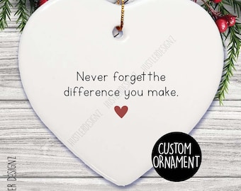 Pode incluir: Um ornamento de cerâmica branco em forma de coração com o texto preto Never forget the difference you make. e um pequeno ícone de coração vermelho. O ornamento inclui um cordão dourado e é exibido sobre um fundo de madeira rústica.