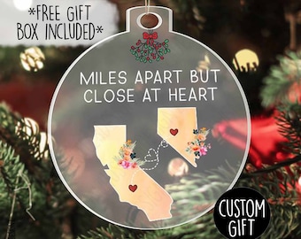 Puede incluir: Adorno navideño de acrílico transparente con el texto «MILES APART BUT CLOSE AT HEART». El adorno presenta dos contornos de estados, cada uno con un corazón y detalles florales. También se ve una ramita de muérdago y las palabras «CUSTOM GIFT».