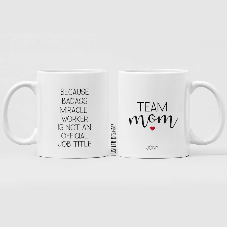 May include: Two white ceramic mugs with black text. One mug says "Because badass miracle worker is not an official job title". The other mug says "Team mom" with a small red heart and the name "Jony" underneath.