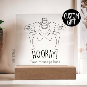 May include: A clear acrylic sign with a black line drawing of a skeleton's hips with the word "HIP" in speech bubbles. The sign reads "HOORAY! Your message here" and has a wooden base. The sign has a sticker that says "CUSTOM GIFT".