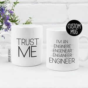 Könnte beinhalten: Zwei weiße Keramikbecher mit schwarzem Text. Der Becher links sagt "TRUST ME". Der Becher rechts sagt "I'M AN ENGINEER" wobei das Wort "ENGINEER" in den Zeilen darüber falsch geschrieben ist. Der Text "CUSTOM MUG" befindet sich in einem Kreis über dem Becher rechts.