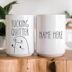 Puede incluir: Dos tazas de cerámica blancas. Una taza tiene el texto "FUCKING QUITTER" con una figura de dibujos animados haciendo una peineta. La otra taza tiene el texto "NAME HERE". Ambas tazas tienen un asa blanca.