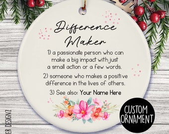 May include: A white ceramic ornament with the text "Difference Maker" and definitions. It features a floral design and the words "Custom Ornament". The ornament is hanging with a gold string, with greenery and red berries in the background.