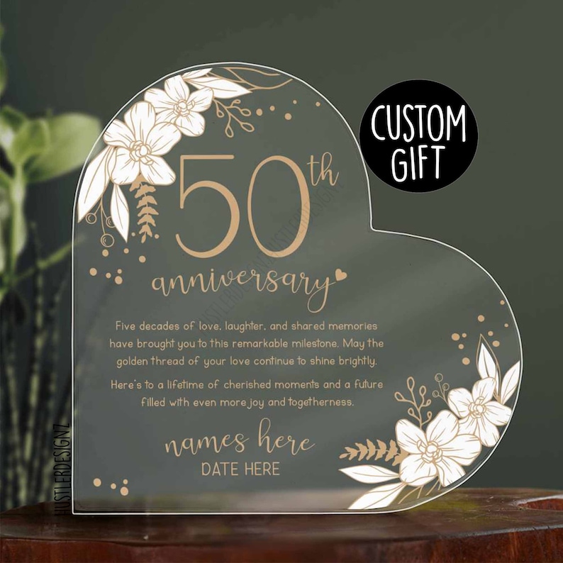 Puede incluir: Una placa de acr&iacute;lico transparente en forma de coraz&oacute;n con un dise&ntilde;o de 50 aniversario en oro. La placa presenta un dise&ntilde;o floral con flores blancas y hojas doradas. El texto "Cinco d&eacute;cadas de amor, risa y recuerdos compartidos te han tra&iacute;do a este hito extraordinario. Que el hilo dorado de tu amor siga brillando. Aqu&iacute; tienes una vida de momentos preciados y un futuro lleno de a&uacute;n m&aacute;s alegr&iacute;a y uni&oacute;n. nombres aqu&iacute; FECHA AQU&Iacute;" est&aacute; impreso en oro en la placa.