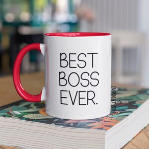 May include: White ceramic mug with a red rim and handle. The mug has the text "BEST BOSS EVER." printed in black.