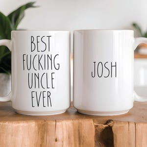 May include: Two white ceramic mugs. One mug has the text "BEST FUCKING UNCLE EVER" in black, the other mug has the name "JOSH" in black. Both mugs have a white handle and are on a wooden surface.