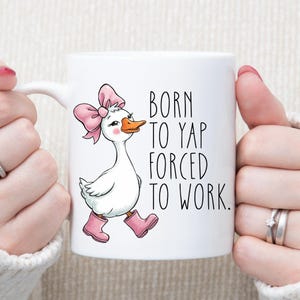 Puede incluir: Taza de cerámica blanca con un ganso de dibujos animados con un lazo rosa y botas, y el texto "BORN TO YAP FORCED TO WORK". El ganso tiene un pico naranja. La taza tiene un asa blanca.