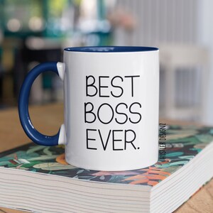 May include: White ceramic coffee mug with a blue rim and handle. The mug has black text that reads "BEST BOSS EVER." The text "HUSTLER DESIGNZ" is printed in smaller font on the right side of the mug.