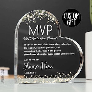 May include: A clear heart-shaped acrylic award with white flowers embedded inside. The text reads "MVP Most Valuable Parent" and includes a personalized message: "The heart and soul of the team, always cheering the loudest, organizing the best, and supporting the hardest. A one-person powerhouse who makes every season unforgettable. Also known as: Name Here Love, Name"
