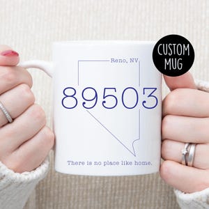 Peut inclure: Une tasse en céramique blanche avec un contour bleu de l'état du Nevada et le texte "Reno, NV" et "89503". La tasse a également le texte "There is no place like home."