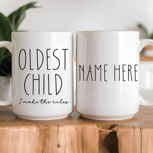 May include: Two white ceramic mugs with black text. One mug says "Oldest Child I make the rules." The other mug says "Name Here".