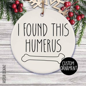 May include: A white ceramic Christmas ornament with the text "I FOUND THIS HUMERUS" in black. A bone illustration is below the text. A black circle with "CUSTOM ORNAMENT" is on the bottom right.