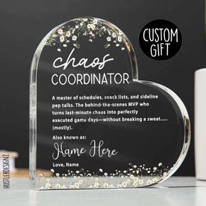 Könnte beinhalten: Ein durchsichtiger, herzförmiger Acryl-Briefbeschwerer mit weißem Blumendesign und dem Text "Chaos-Koordinator" mit einer Definition. Der Text "Custom Gift" befindet sich in der oberen rechten Ecke. Der Text "Auch bekannt als: Name Here" befindet sich am unteren Rand des Briefbeschwerers.