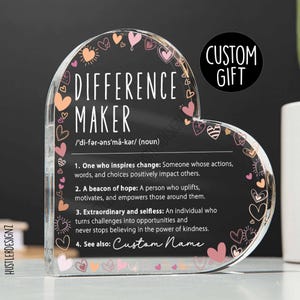 Pode incluir: Placa de acrílico em forma de coração com as palavras "DIFFERENCE MAKER" e sua definição. A placa tem um fundo preto com corações rosa e pêssego. O texto "CUSTOM GIFT" está em um círculo.