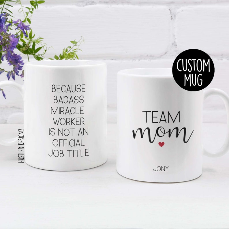 May include: Two white ceramic mugs with black text. One mug says "Because Badass Miracle Worker is not an Official Job Title". The other mug says "Team Mom" with a red heart and the name "Jony" below.
