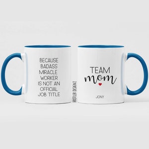 May include: Two white coffee mugs with blue rims. One mug says "Because badass miracle worker is not an official job title". The other mug says "Team mom" with a red heart and the name "Jony".