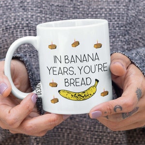 May include: White ceramic mug with a black and yellow banana illustration and the text "In banana years, you're bread". There are small brown squares with a yellow candle on top scattered around the banana.
